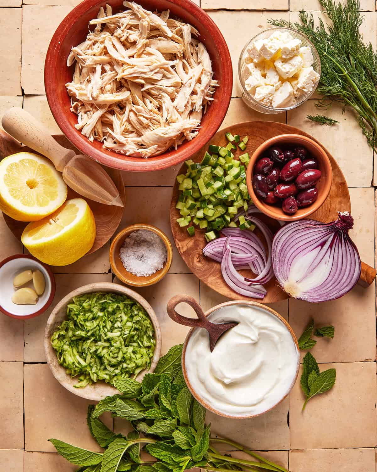 A top-down view of ingredients on a tiled surface, including shredded chicken, feta, olives, chopped celery, red onion, Greek yogurt, grated cucumber, lemon halves, garlic cloves, fresh dill, mint, and a small bowl of salt.
