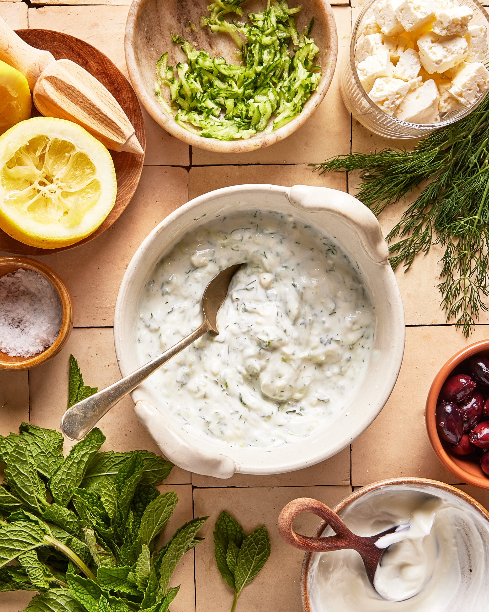 A bowl of tzatziki sauce with a spoon, surrounded by ingredients: grated cucumber, feta cheese, lemon halves, dill, mint, salt, olives, and yogurt, all arranged on a tiled surface.