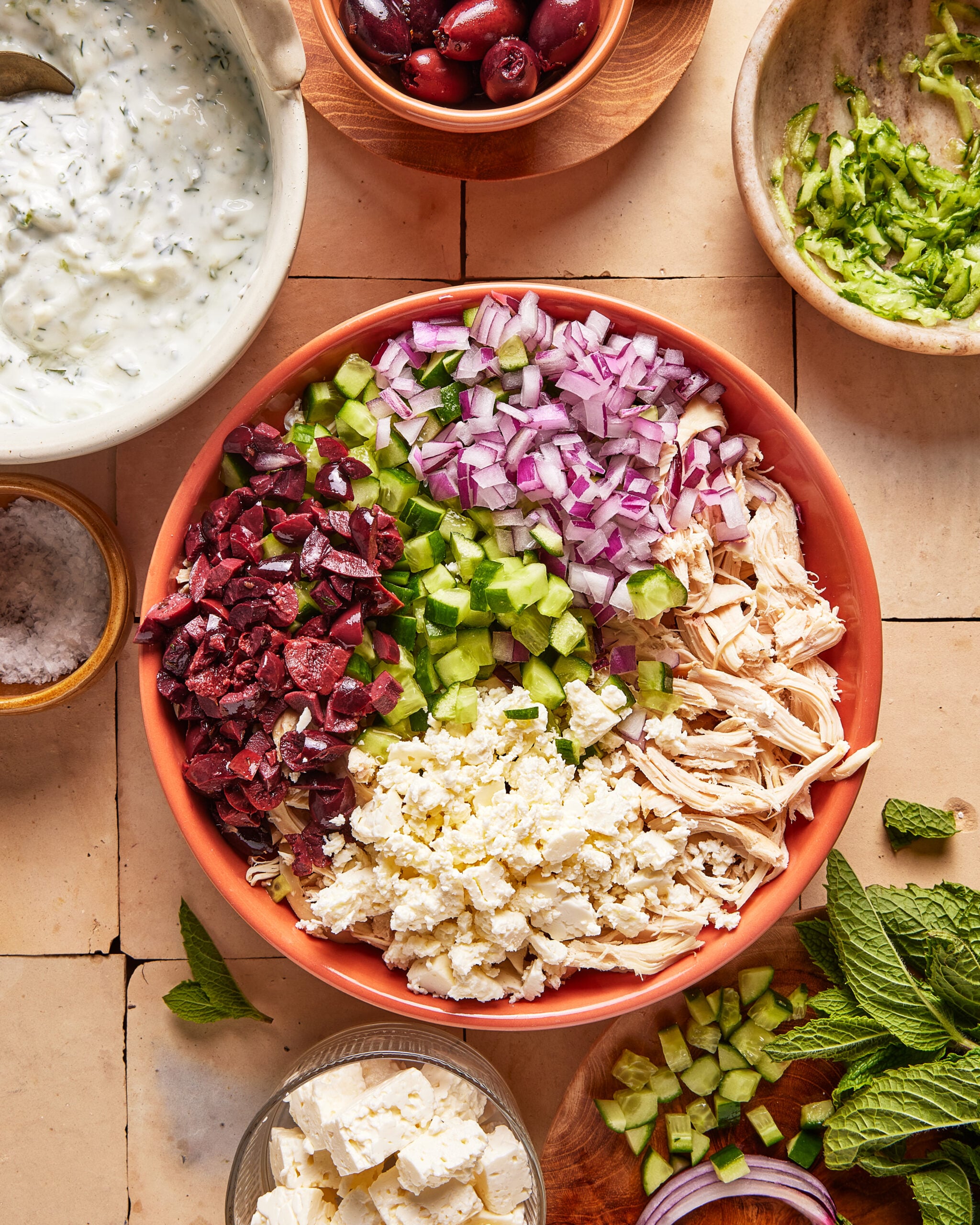 A bowl filled with chopped red onions, cucumbers, Kalamata olives, shredded chicken, and crumbled feta cheese. Surrounding bowls hold tzatziki, olives, feta, shredded cucumber, salt, and fresh mint leaves.