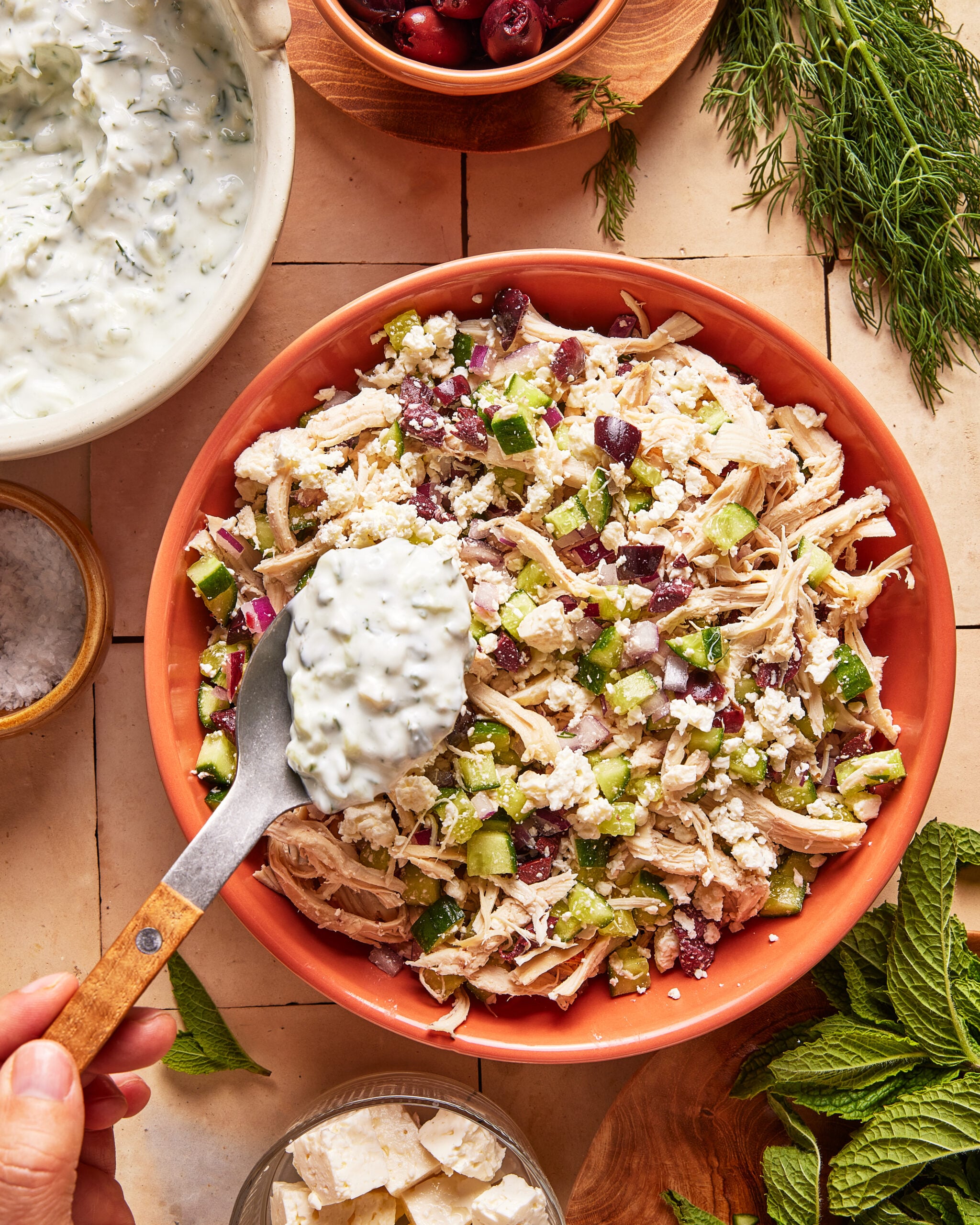 A hand holds a spoon of creamy tzatziki sauce above a bowl of shredded chicken salad with cucumbers, olives, feta, and herbs. Surrounding the bowl are fresh herbs, feta, salt, and a bowl of tzatziki.