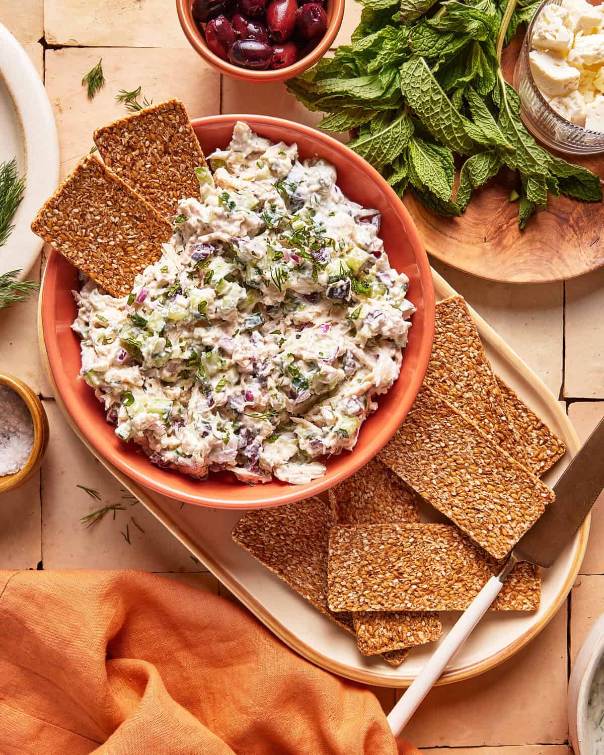 A bowl of creamy salad with herbs, capers, and onions is surrounded by rectangular seeded crackers. Fresh mint, a bowl of olives, salt, and a knife are nearby, arranged on a tiled surface with an orange napkin.