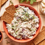 A bowl of creamy chicken salad with herbs and chopped vegetables, garnished with fresh dill, surrounded by rectangular seed crackers on a tiled surface. Mint leaves and cheese are visible in the background.