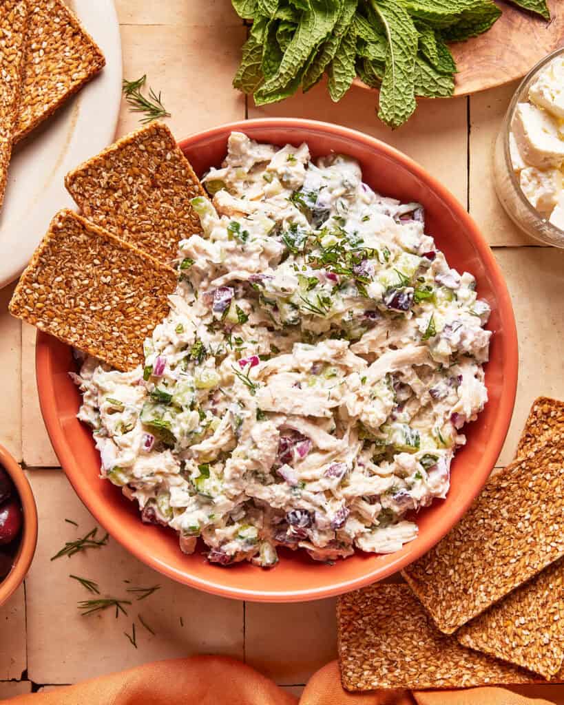 A bowl of creamy chicken salad with herbs and chopped vegetables, garnished with fresh dill, surrounded by rectangular seed crackers on a tiled surface. Mint leaves and cheese are visible in the background.