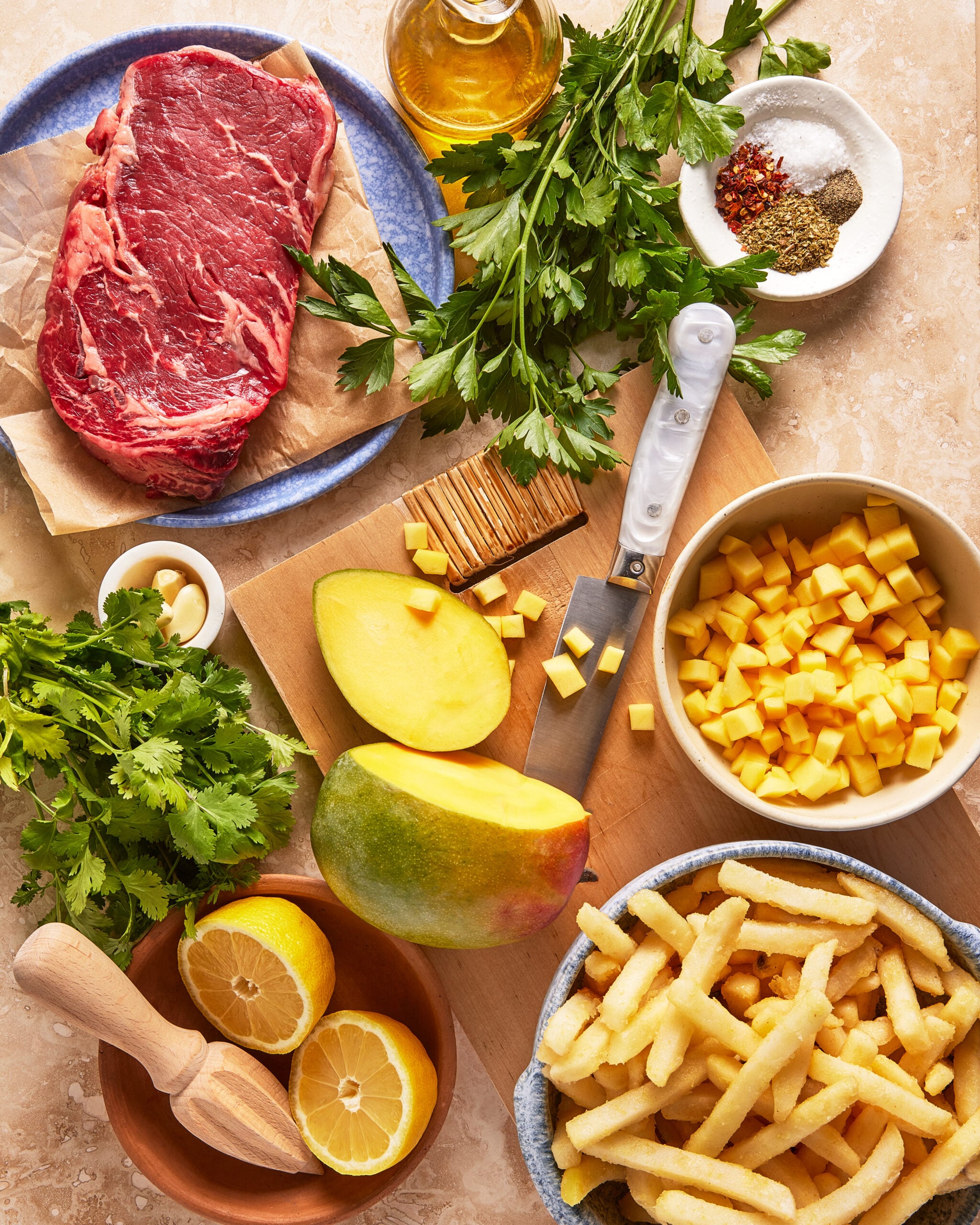 A flat lay of ingredients including a raw steak, fresh herbs, olive oil, spices, a mango (whole and diced), lemon halves, French fries, and a knife on a cutting board.