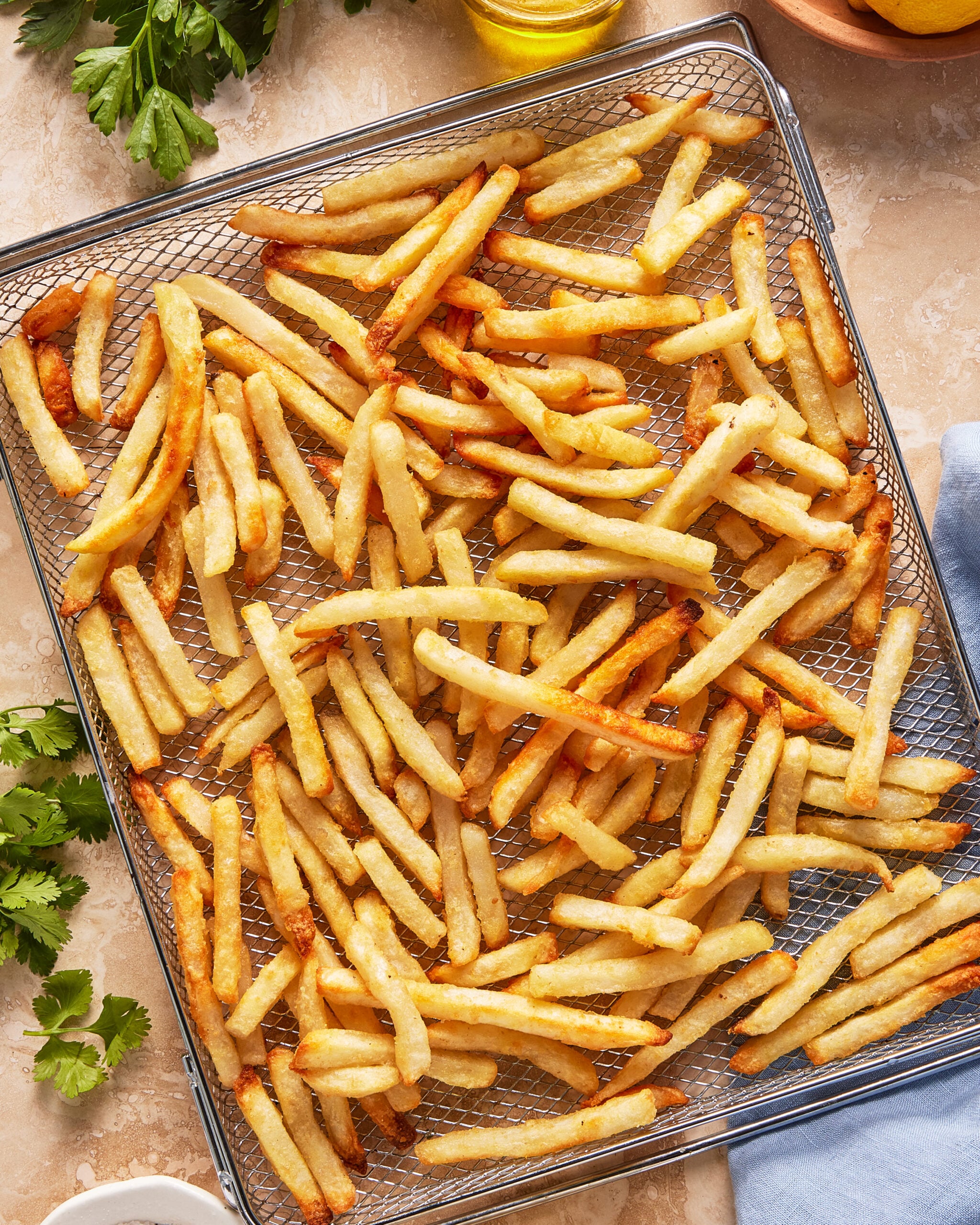 A wire rack filled with golden, crispy French fries, surrounded by fresh parsley and a blue napkin on a light-colored surface.