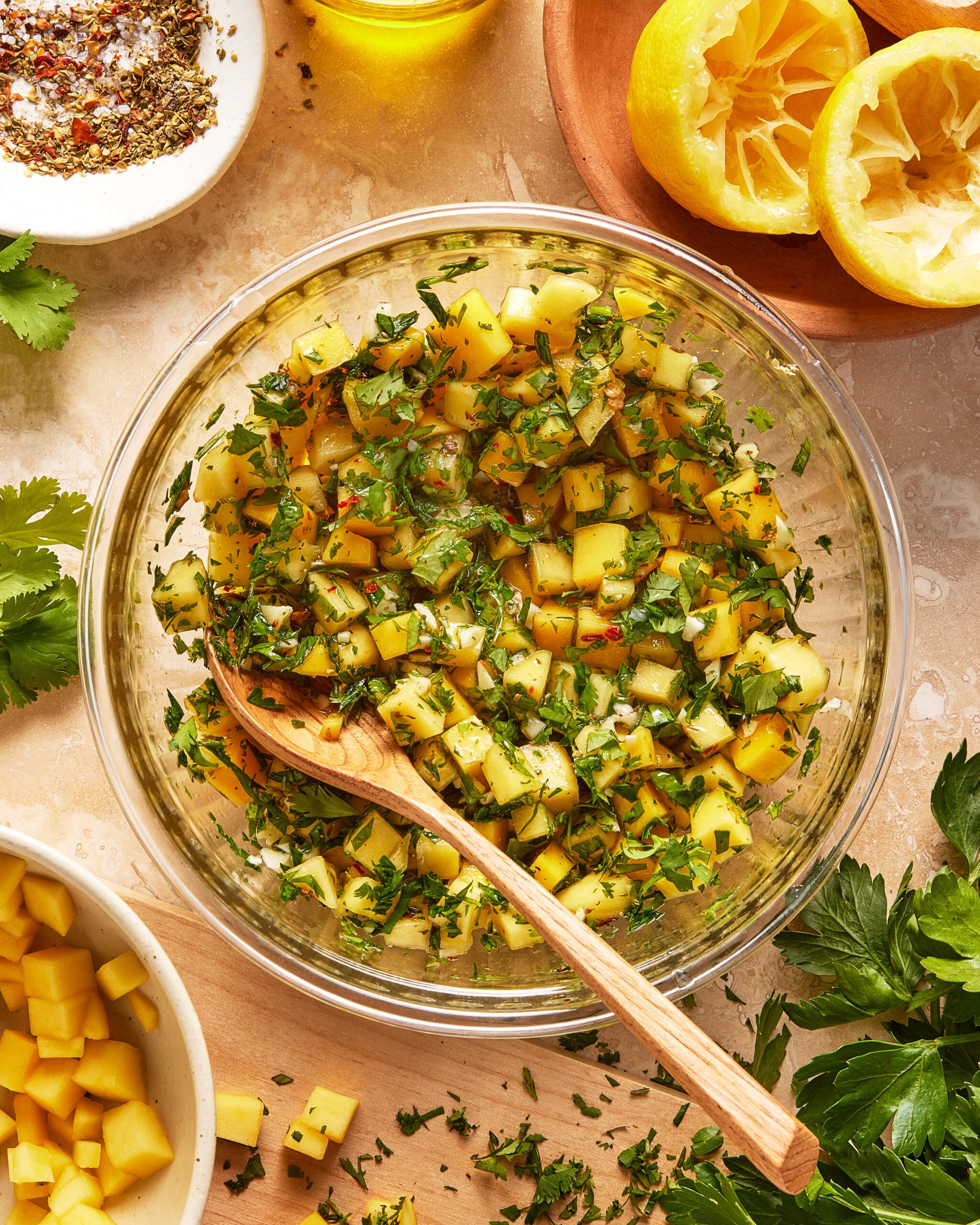 A glass bowl filled with a mango salsa mixture, including diced mango, chopped herbs, and spices, with a wooden spoon. Around the bowl are squeezed lemons, fresh herbs, and a bowl of diced mango.