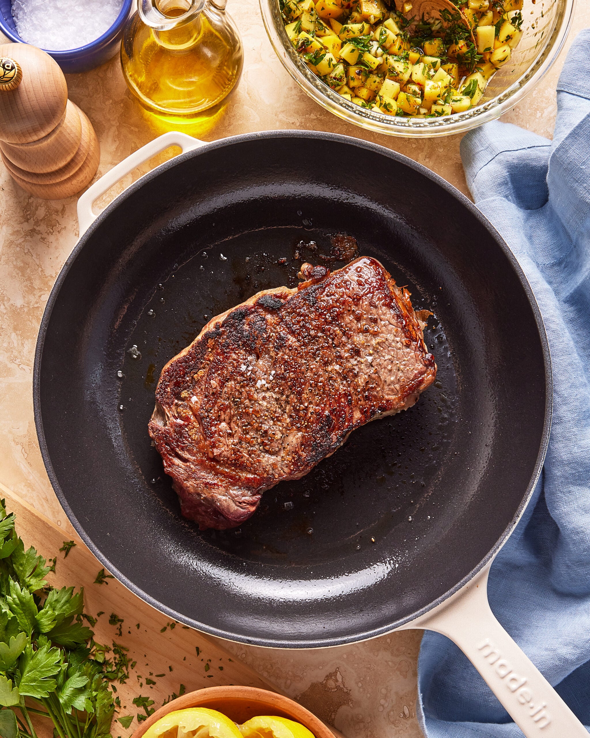 A cooked steak sits in a black skillet on a beige countertop, surrounded by a bowl of chopped herbs and potatoes, a bottle of olive oil, a pepper grinder, parsley, lemon slices, and a blue cloth napkin.