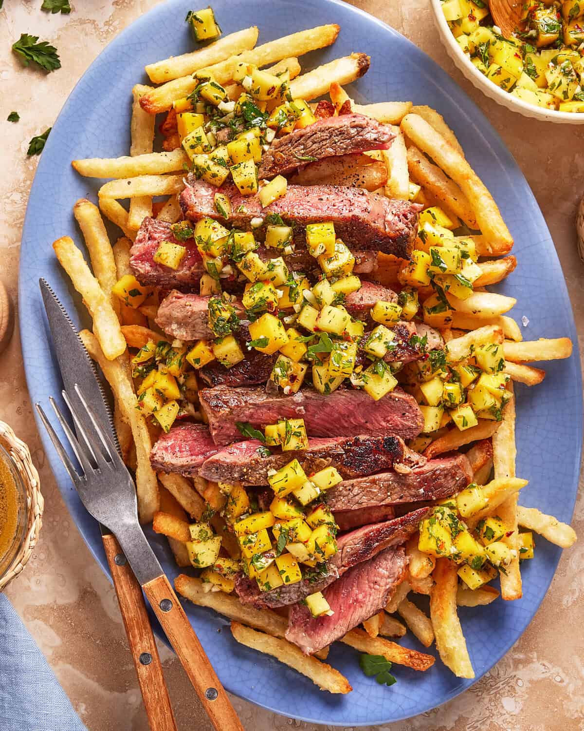 Sliced grilled steak served on top of golden French fries, garnished with a fresh pineapple salsa, on a blue platter with a fork and knife beside it.