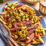 A plate of French fries topped with sliced grilled steak and a chunky mango salsa, served with utensils. Extra mango salsa is in small bowls nearby, and the table is decorated with drinks and herbs.