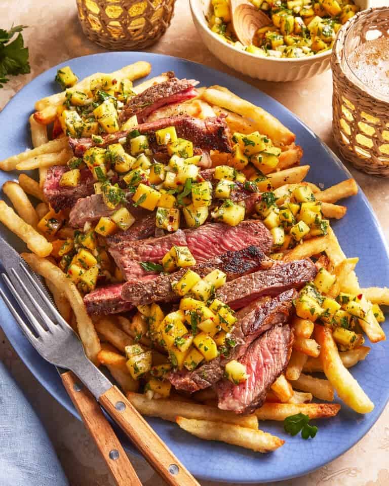 A plate of French fries topped with sliced grilled steak and a chunky mango salsa, served with utensils. Extra mango salsa is in small bowls nearby, and the table is decorated with drinks and herbs.