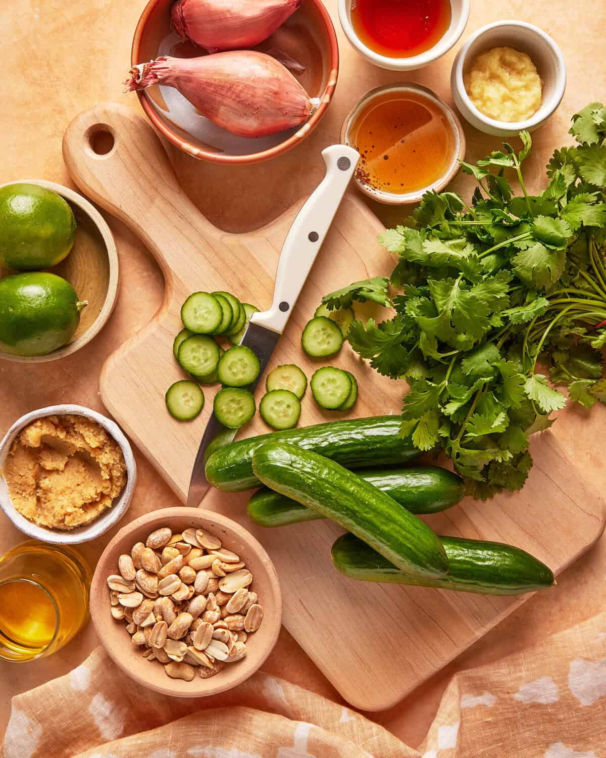 A cutting board with sliced cucumbers, whole cucumbers, cilantro, and a knife, surrounded by bowls of peanuts, brown sugar, oil, lime, shallots, honey, and minced ginger on a light surface.