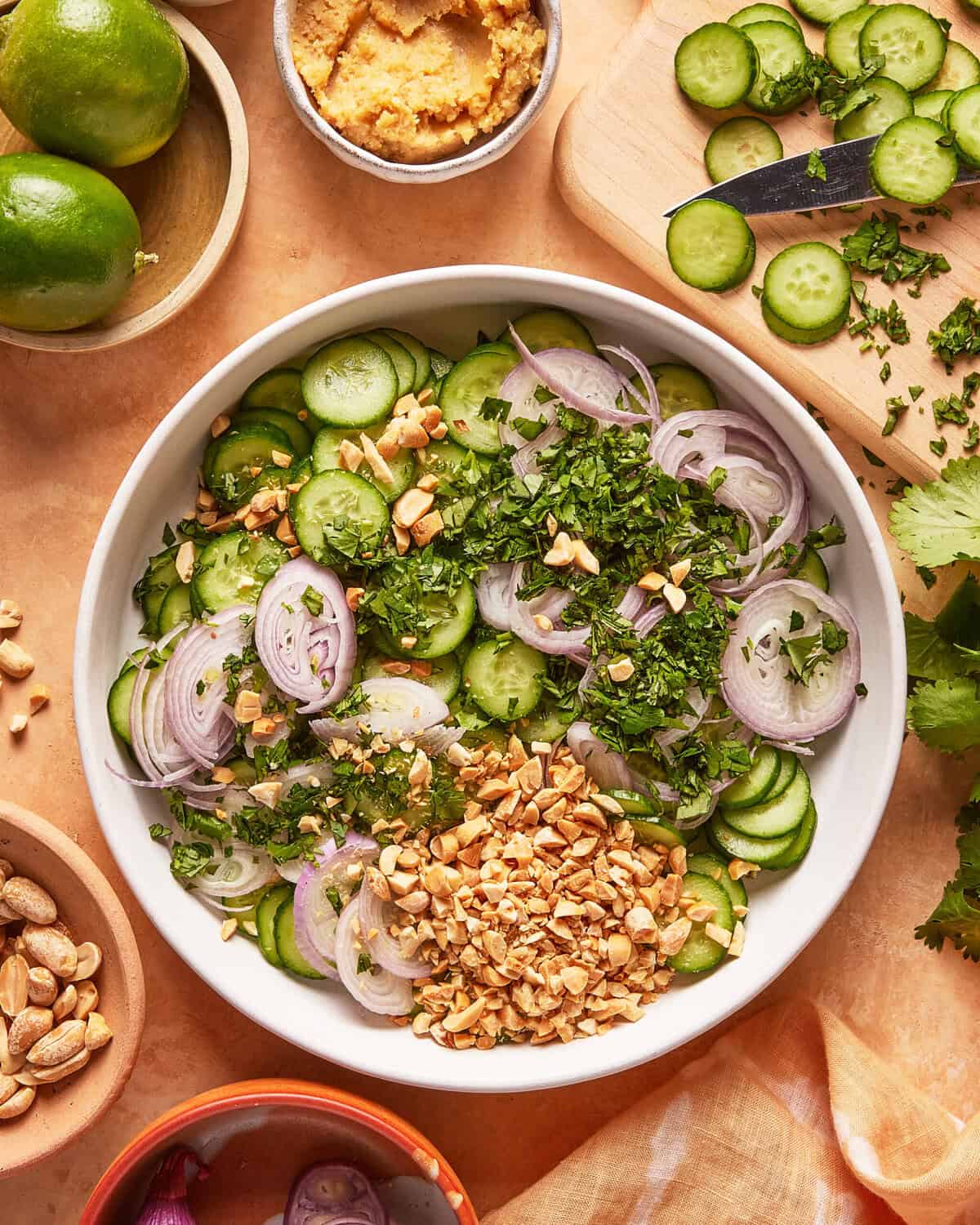 A bowl of cucumber salad with sliced cucumbers, shallots, chopped herbs, and crushed peanuts. Nearby are a cutting board with cucumber slices, a knife, limes, fresh cilantro, and bowls of peanuts and dressing.