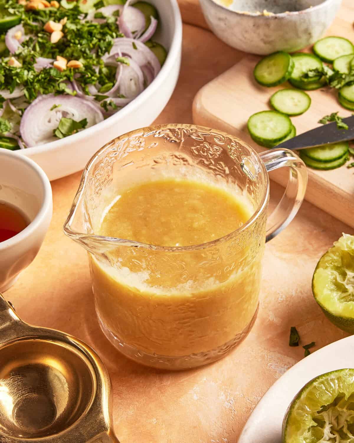 A glass pitcher filled with golden salad dressing sits on a table surrounded by sliced cucumbers, fresh lime halves, herbs, sliced onions, and a salad bowl. A knife and small bowl of dressing are nearby.
