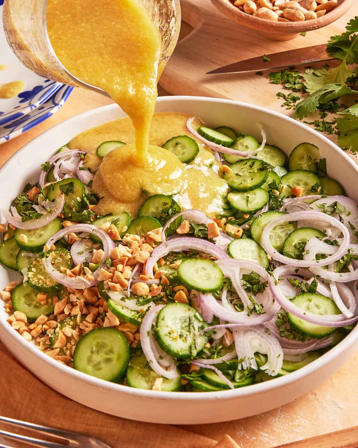 A hand pours yellow dressing over a salad with sliced cucumbers, red onions, chopped peanuts, and fresh herbs in a white bowl. Other ingredients and utensils are visible in the background.