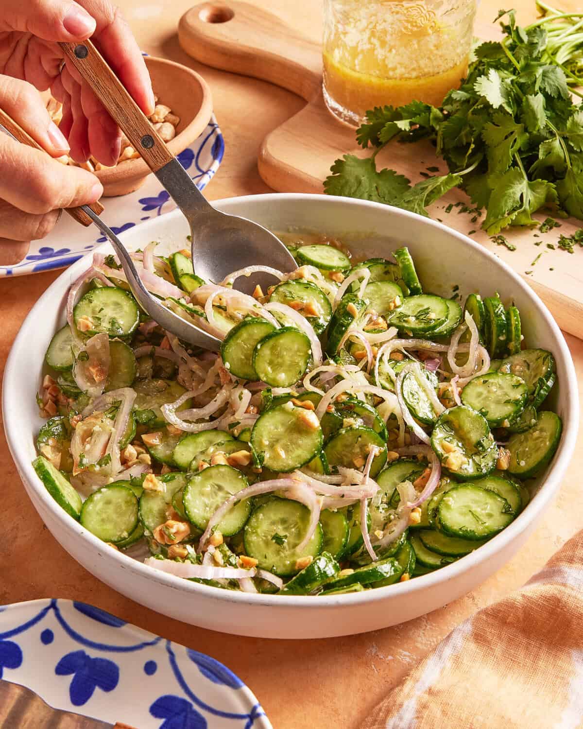 A hand serves a fresh cucumber salad with sliced shallots and chopped nuts from a white bowl. Fresh cilantro, a jar of dressing, and a wooden cutting board are nearby on a light-colored countertop.