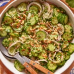 A bowl of cucumber salad with sliced cucumbers, red onions, chopped herbs, crushed nuts, and red pepper flakes, served with wooden-handled salad tongs.