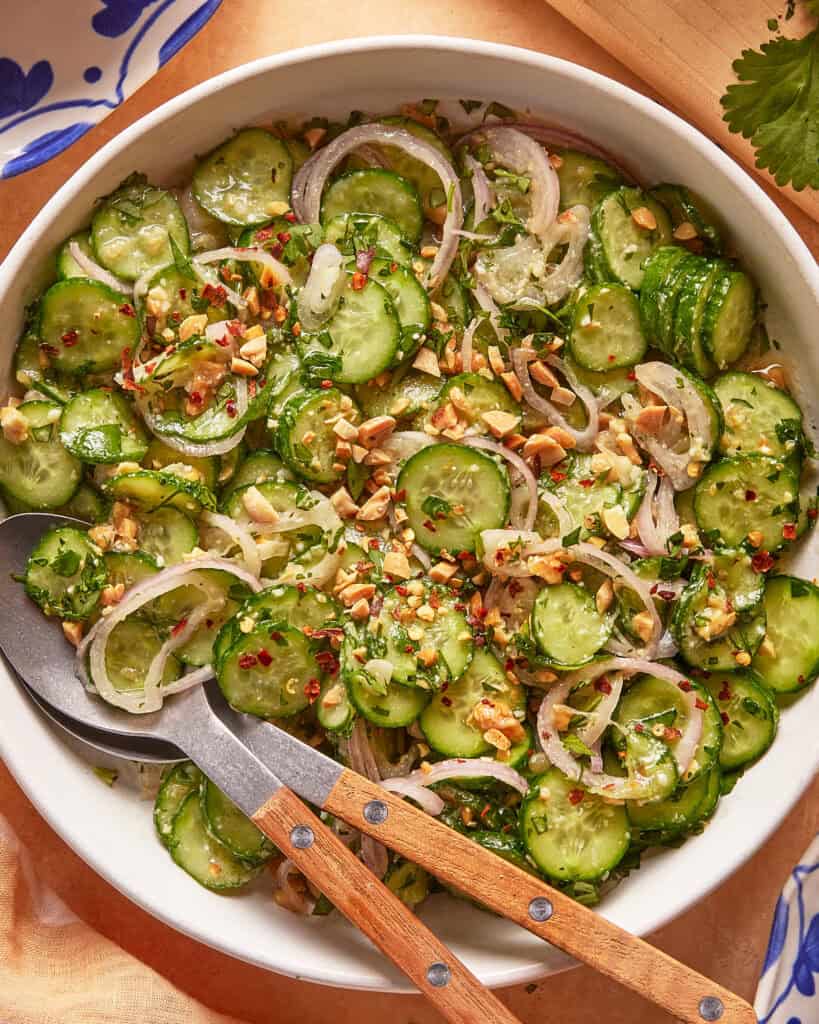 A bowl of cucumber salad with sliced cucumbers, red onions, chopped herbs, crushed nuts, and red pepper flakes, served with wooden-handled salad tongs.