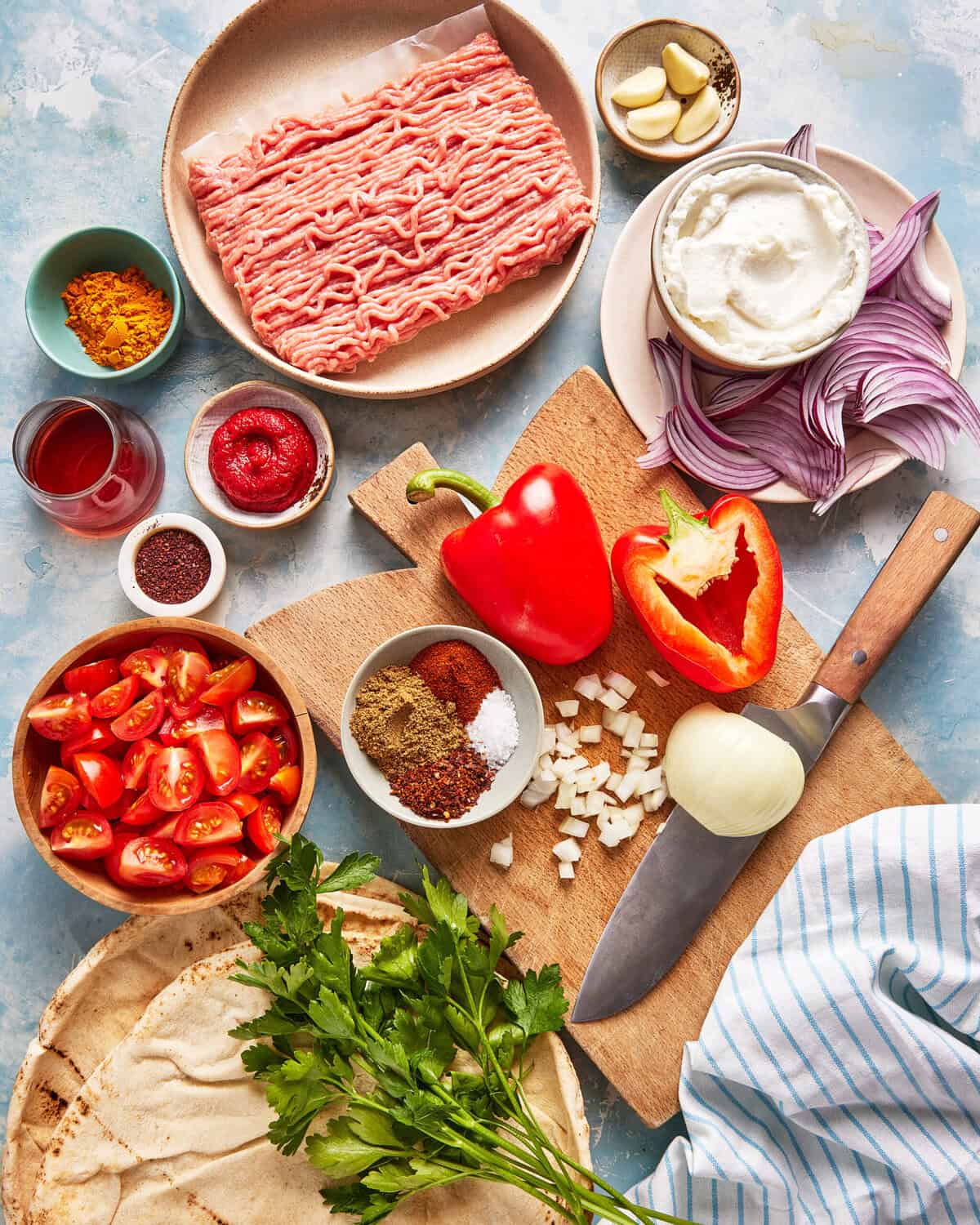 Assorted ingredients on a table, including ground meat, yogurt, sliced red onion, garlic, red pepper, cherry tomatoes, spices, tomato paste, chopped herbs, flatbread, and a knife on a cutting board.