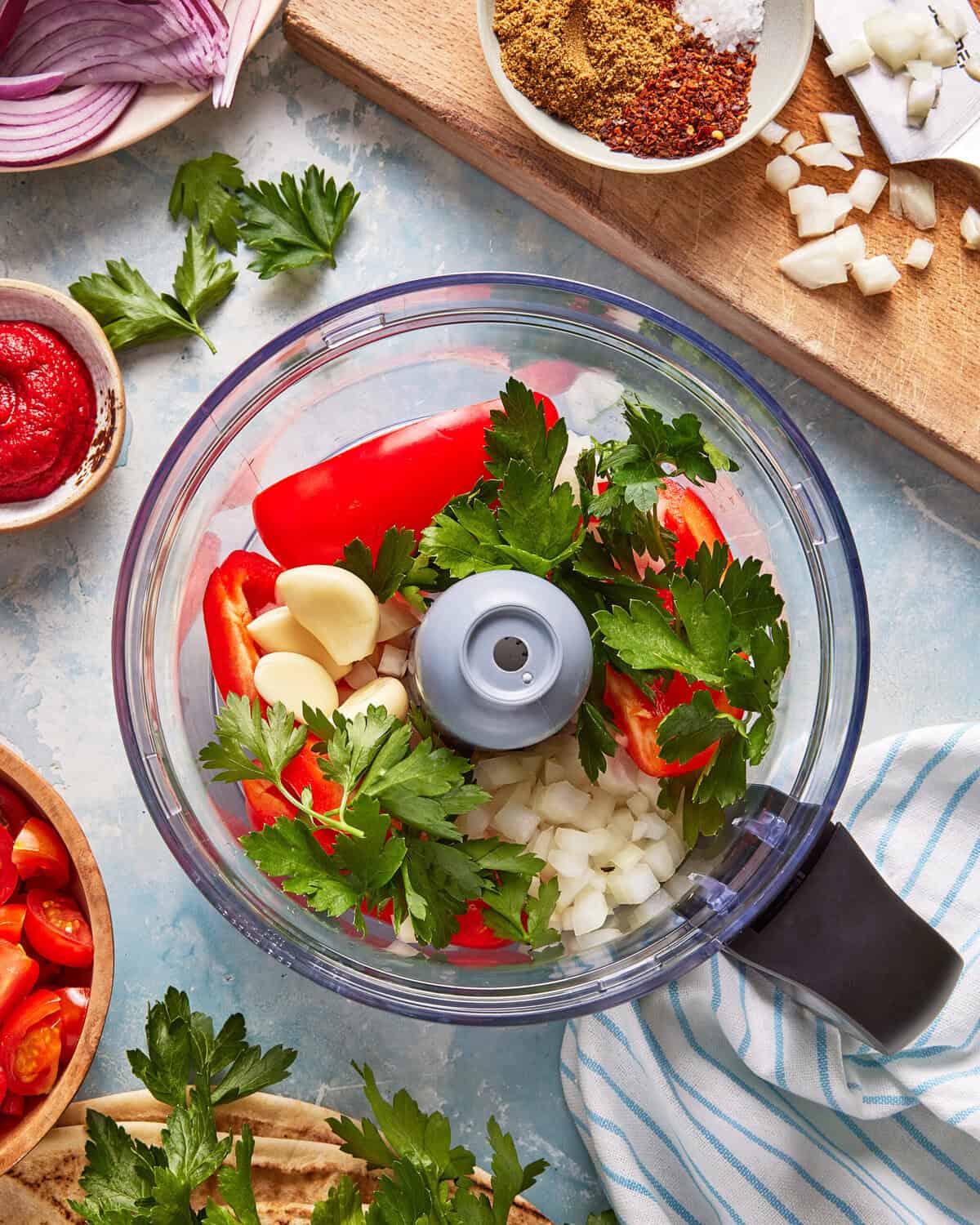 A food processor bowl filled with parsley, garlic cloves, chopped onion, and red bell pepper. Surrounding it are spices, tomato paste, chopped tomatoes, parsley, sliced onion, and a striped cloth.