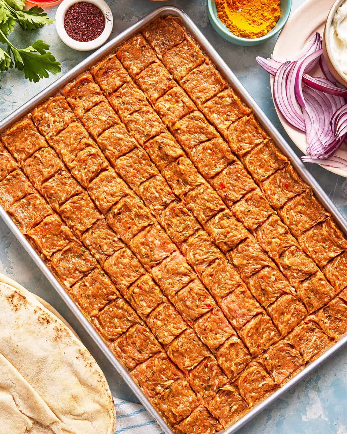 A large metal tray filled with neatly scored, uncooked minced meat mixture. Surrounding the tray are plates with sliced red onions, spices, flatbread, and fresh herbs on a light blue surface.