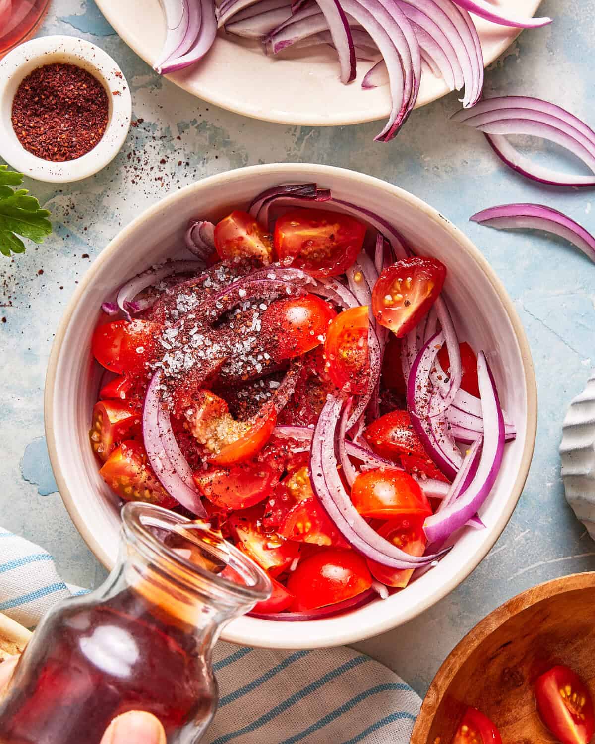 A bowl filled with sliced red onions and chopped tomatoes, sprinkled with salt and spices. A hand pours dressing from a glass bottle. Small bowls of spices and extra onion slices surround the main bowl.