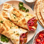 Two pita wraps filled with grilled meat, sliced red onions, and herbs are served on a plate, garnished with chopped parsley. A bowl of sauce, stacked pita bread, and a bowl of tomato salad are nearby.