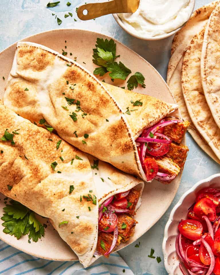 Two pita wraps filled with grilled meat, sliced red onions, and herbs are served on a plate, garnished with chopped parsley. A bowl of sauce, stacked pita bread, and a bowl of tomato salad are nearby.