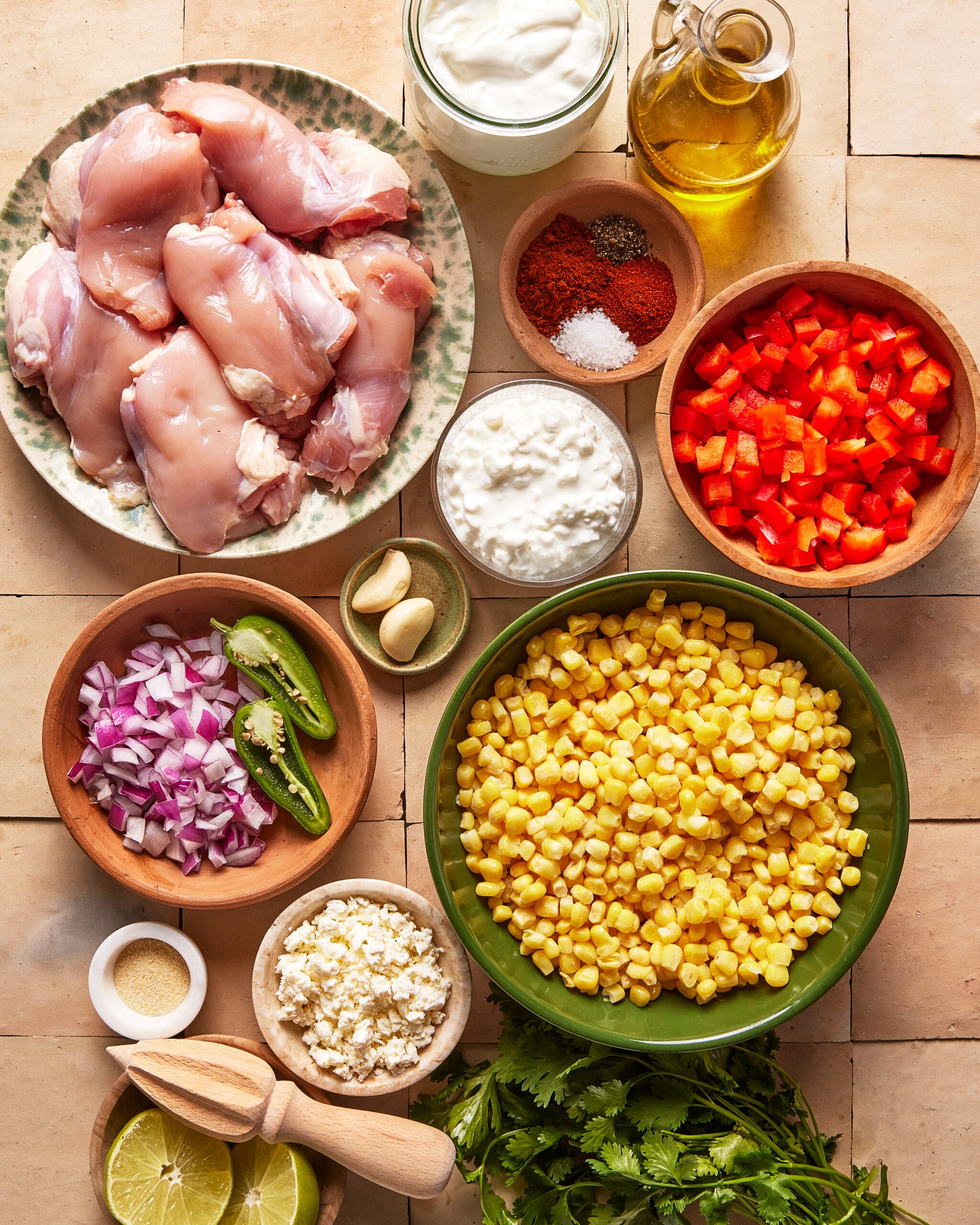 A top-down view of bowls with raw chicken, corn, chopped red bell pepper, red onion, sliced jalapeno, garlic, spices, lime wedges, oil, yogurt, cottage cheese, cheese crumbles, and fresh cilantro on a tiled surface.