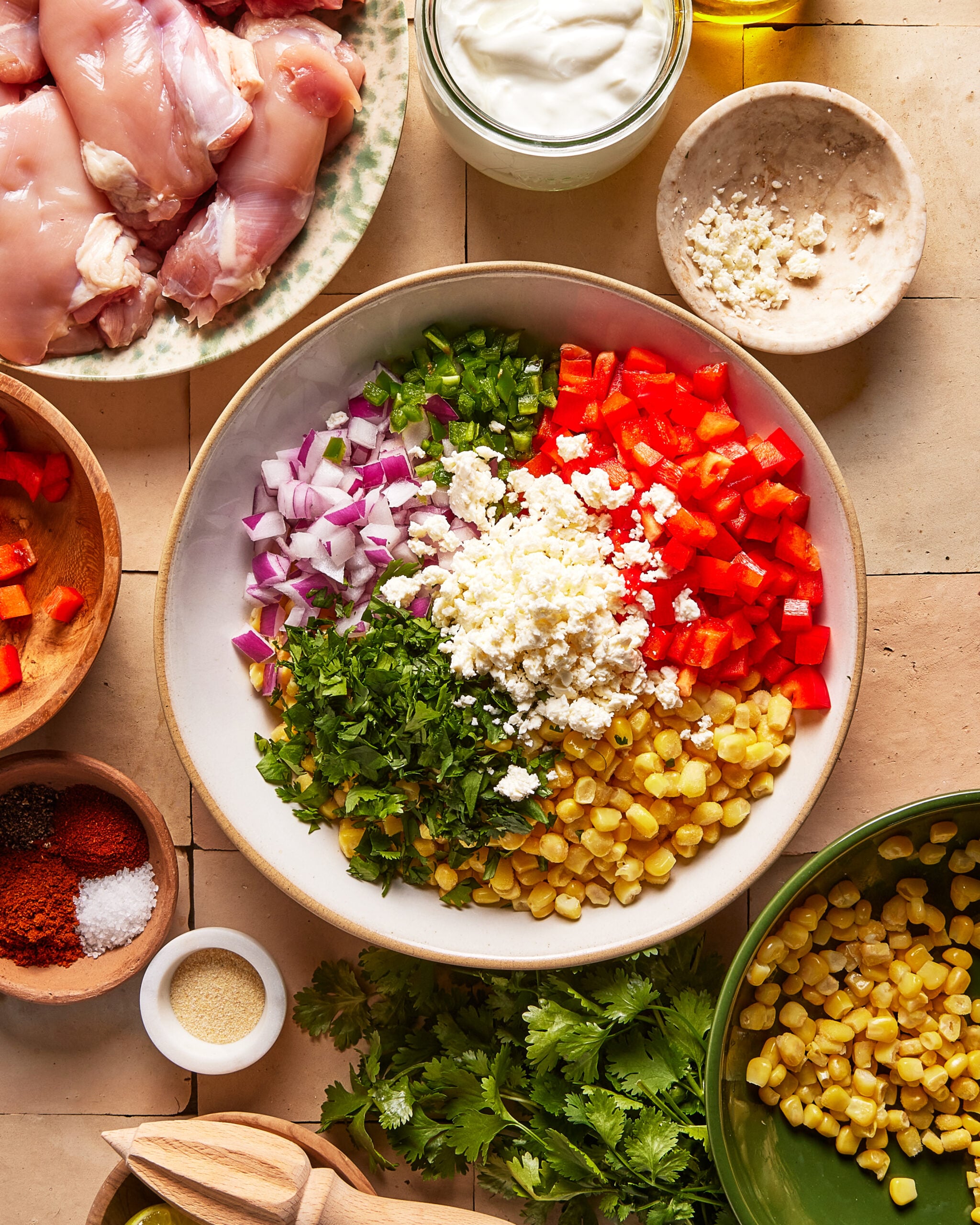 A bowl filled with chopped red bell pepper, corn, cilantro, red onion, jalapeño, and crumbled cheese. Surrounding it are bowls of raw chicken, seasonings, yogurt, cilantro, and olive oil on a tiled surface.