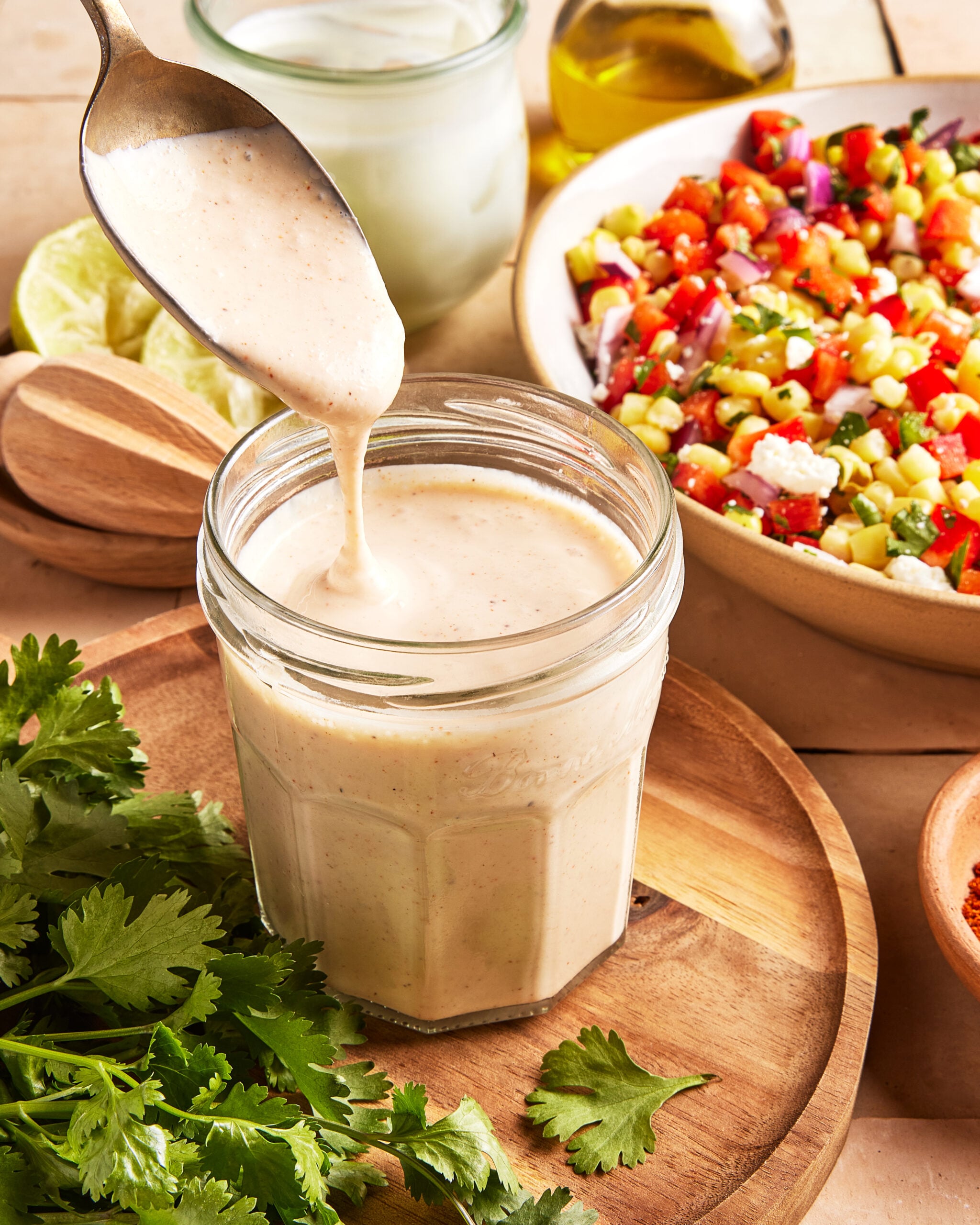 A spoon drizzles creamy dressing into a glass jar on a wooden board, surrounded by fresh cilantro, lime slices, and a colorful vegetable salad in a bowl.
