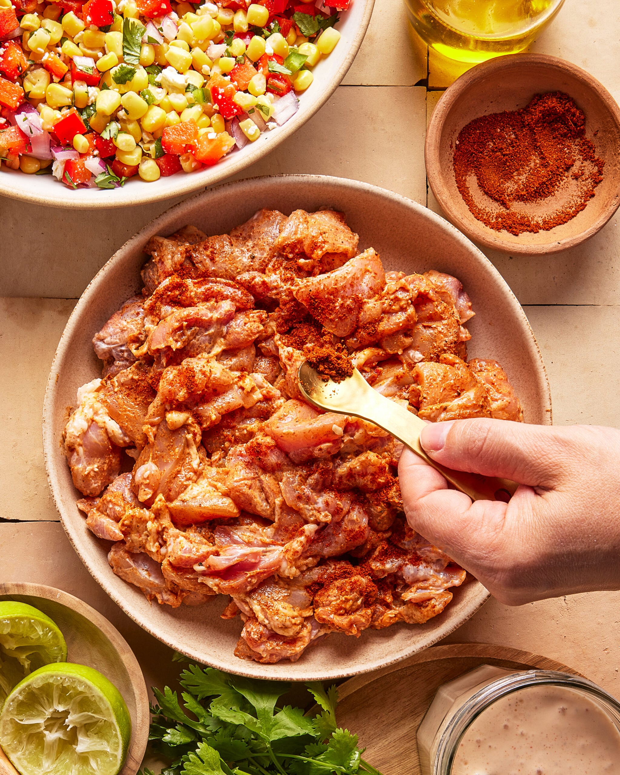 A hand sprinkles spice onto marinated chicken pieces in a bowl, surrounded by a colorful corn salsa, lime halves, fresh cilantro, a small bowl of seasoning, olive oil, and a jar of creamy sauce.