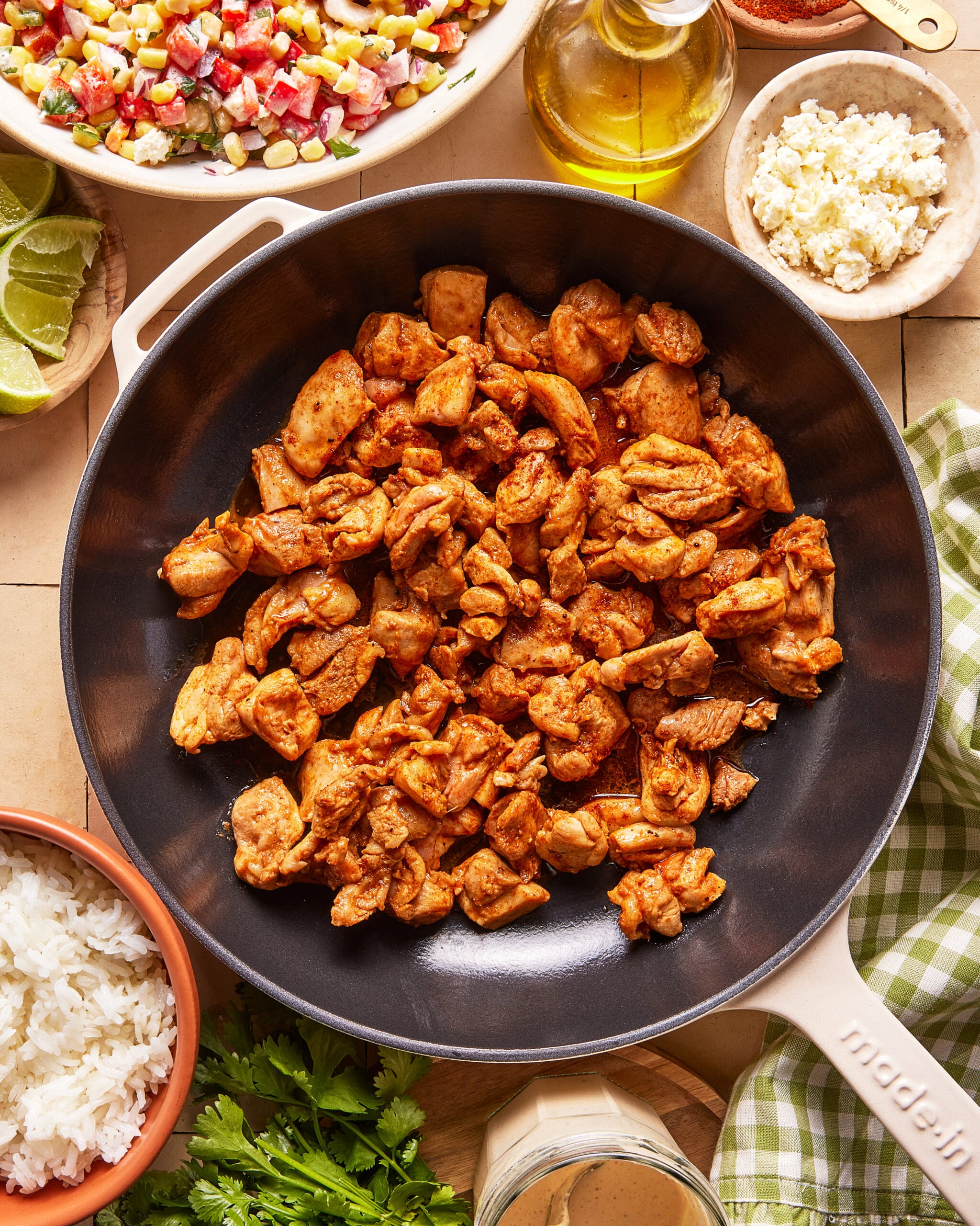 A skillet filled with seasoned, cooked chicken pieces is surrounded by bowls of corn salsa, rice, feta cheese, lime wedges, cilantro, and a bottle of olive oil on a tiled surface.