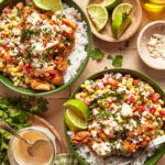 Two green bowls filled with white rice, grilled chicken, corn salsa, crumbled cheese, cilantro, and lime wedges. Small dishes of lime, cheese, and dressing are nearby, along with fresh cilantro and a gingham napkin.
