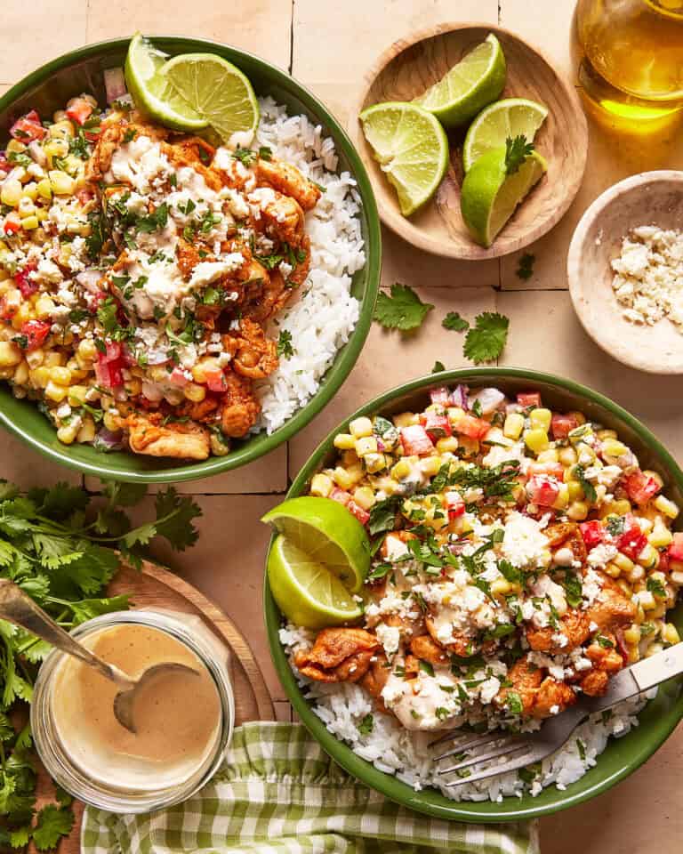Two green bowls filled with white rice, grilled chicken, corn salsa, crumbled cheese, cilantro, and lime wedges. Small dishes of lime, cheese, and dressing are nearby, along with fresh cilantro and a gingham napkin.