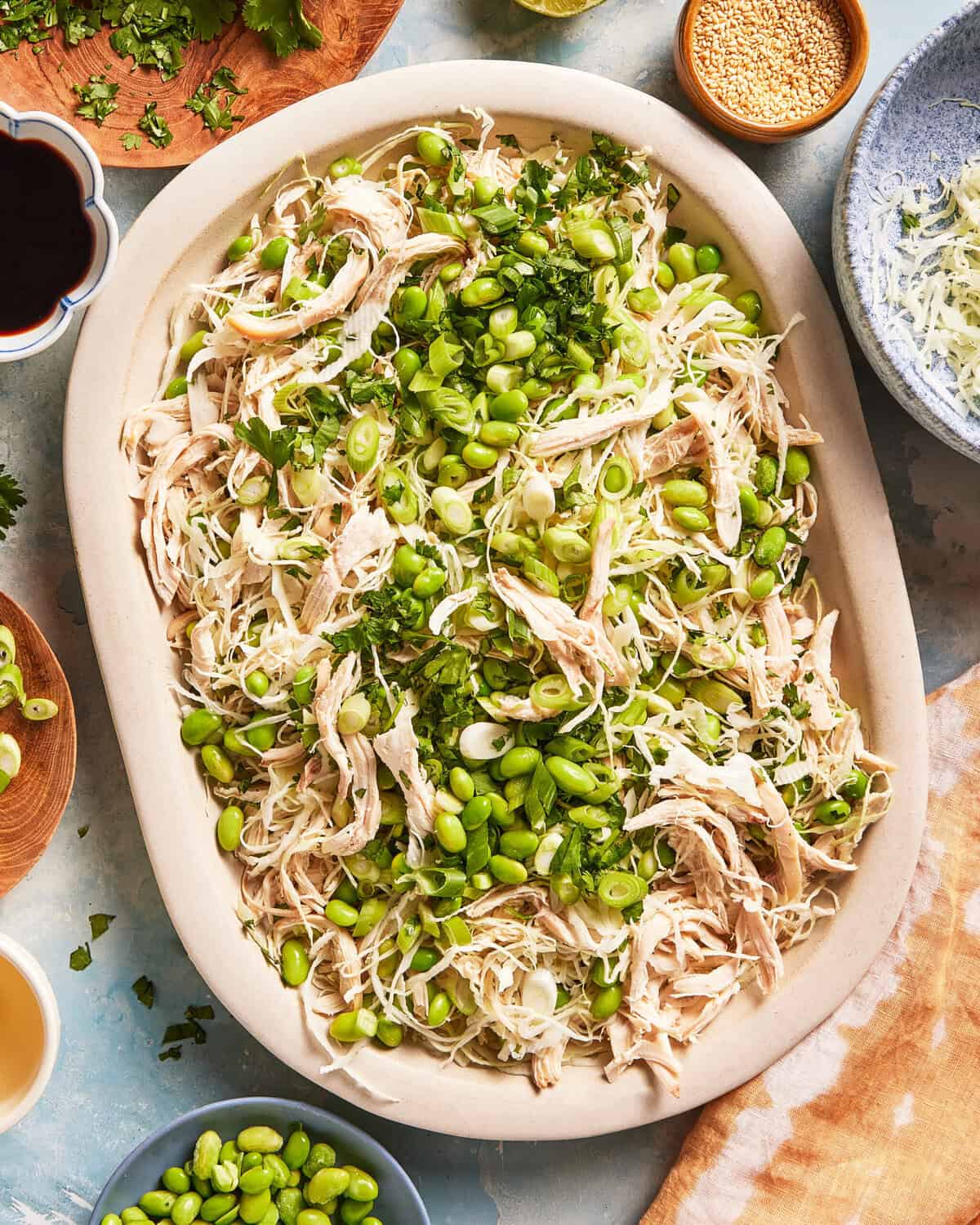 Shredded chicken salad with edamame, shredded cabbage, chopped green onions, and cilantro on a large oval dish, surrounded by small bowls of ingredients.