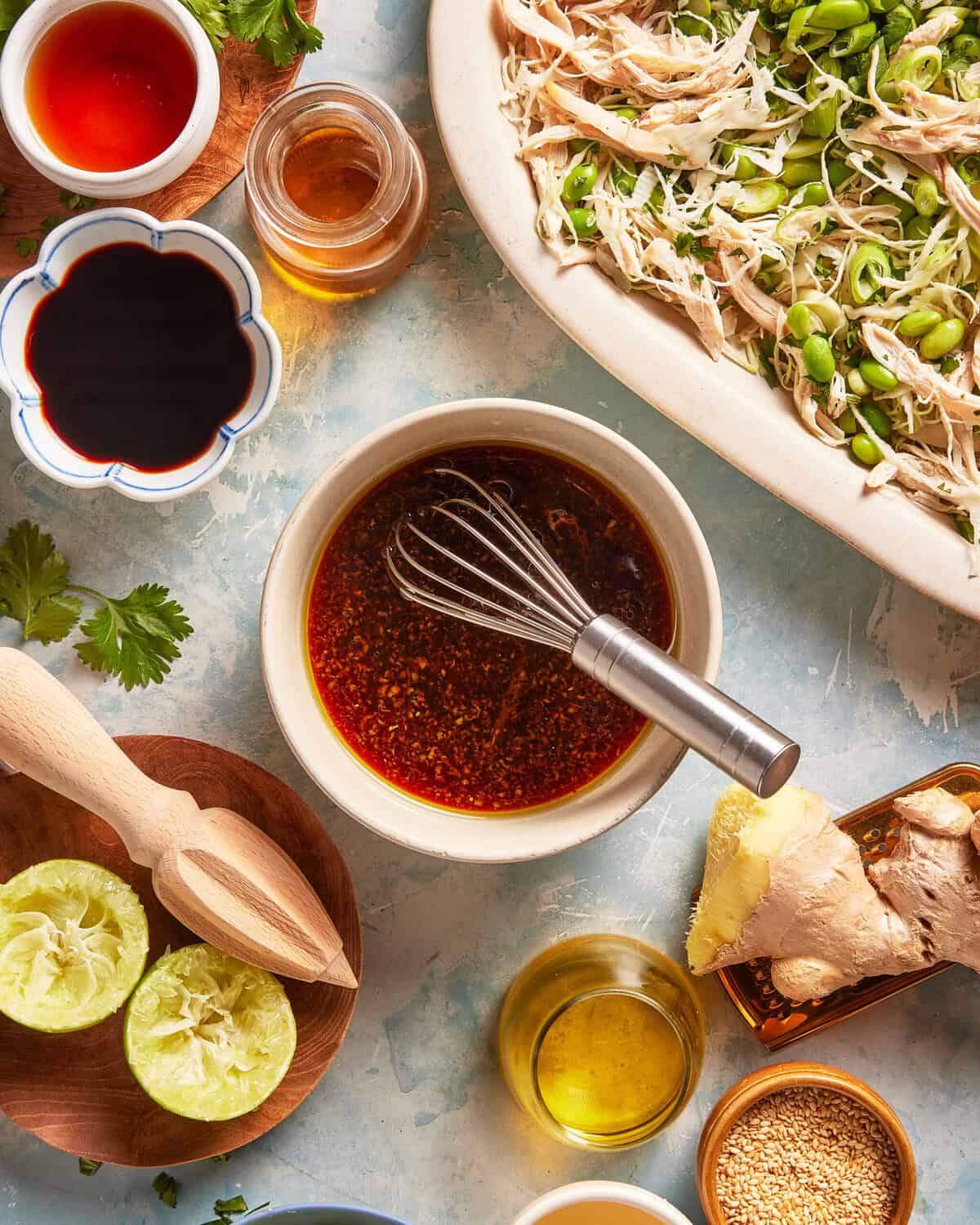 Overhead view of various ingredients, including lime halves, honey, ginger, soy sauce, oil, and a whisk in a bowl with dark liquid, next to a plate of shredded chicken salad.