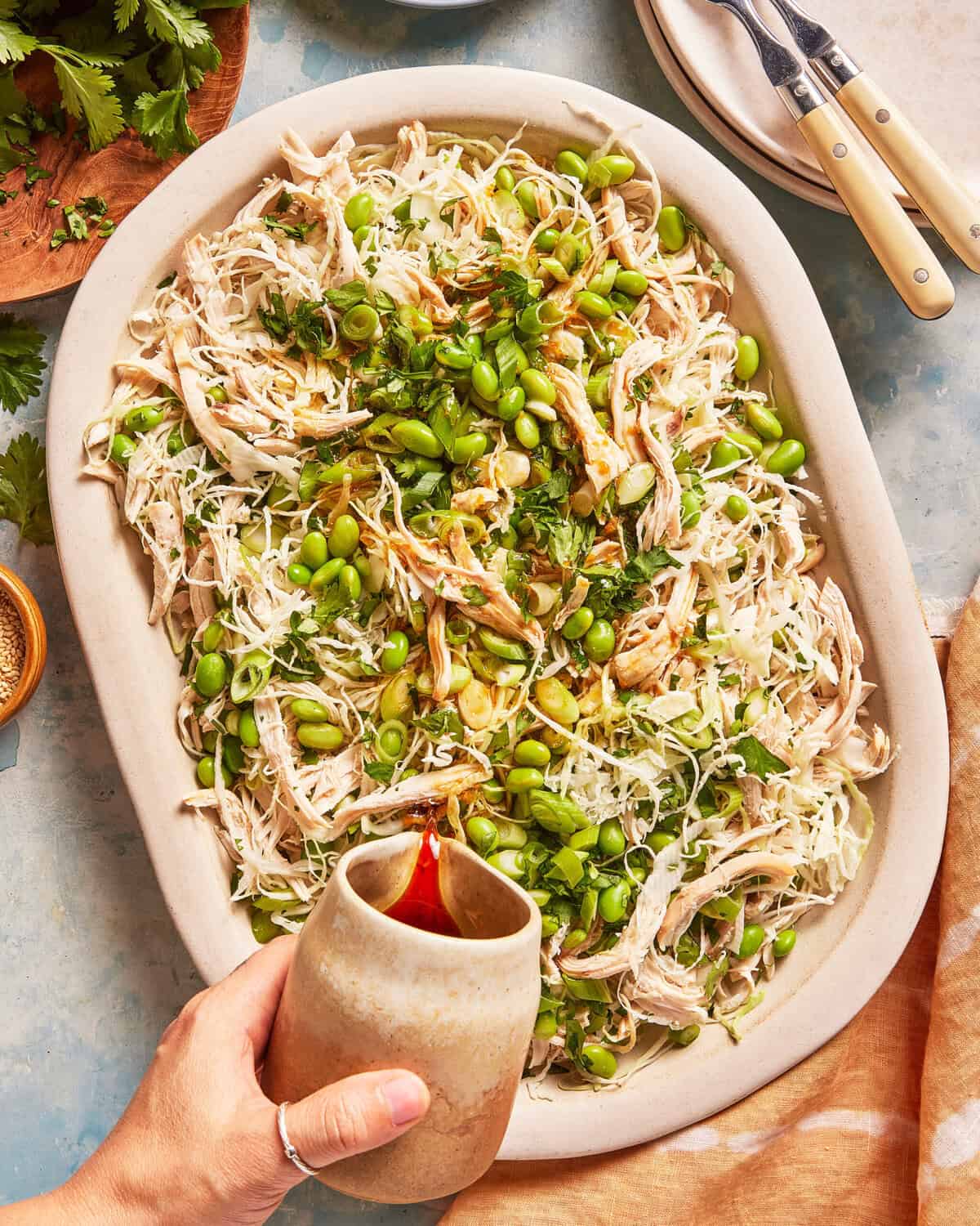 A hand pours dressing over a platter of shredded chicken, edamame, cabbage, and herbs. Plates and utensils are nearby on a light blue surface.