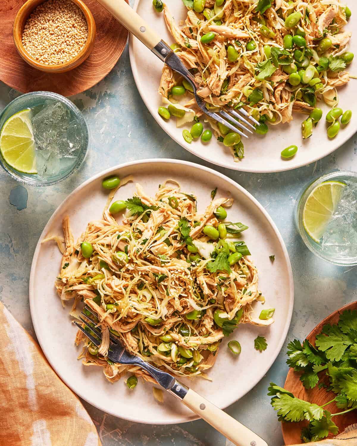 Two plates of shredded chicken salad with edamame, cilantro, and sesame seeds. Lime wedges and glasses of sparkling water are nearby on a light blue surface.