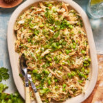 Large oval platter filled with shredded chicken, edamame, chopped scallions, sesame seeds, and herbs. Two serving utensils rest on the edge. Plates, drinks, and fresh cilantro are nearby on a light blue table.