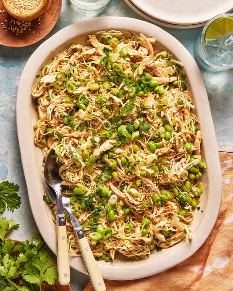 Large oval platter filled with shredded chicken, edamame, chopped scallions, sesame seeds, and herbs. Two serving utensils rest on the edge. Plates, drinks, and fresh cilantro are nearby on a light blue table.