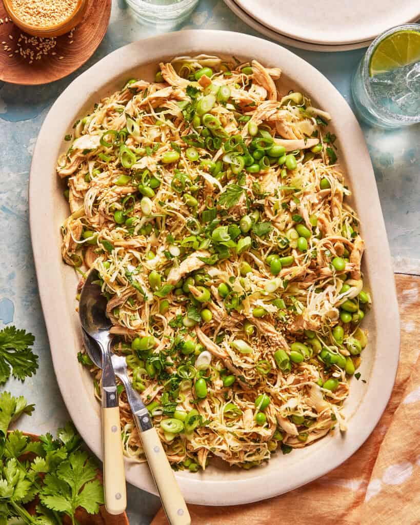 Large oval platter filled with shredded chicken, edamame, chopped scallions, sesame seeds, and herbs. Two serving utensils rest on the edge. Plates, drinks, and fresh cilantro are nearby on a light blue table.
