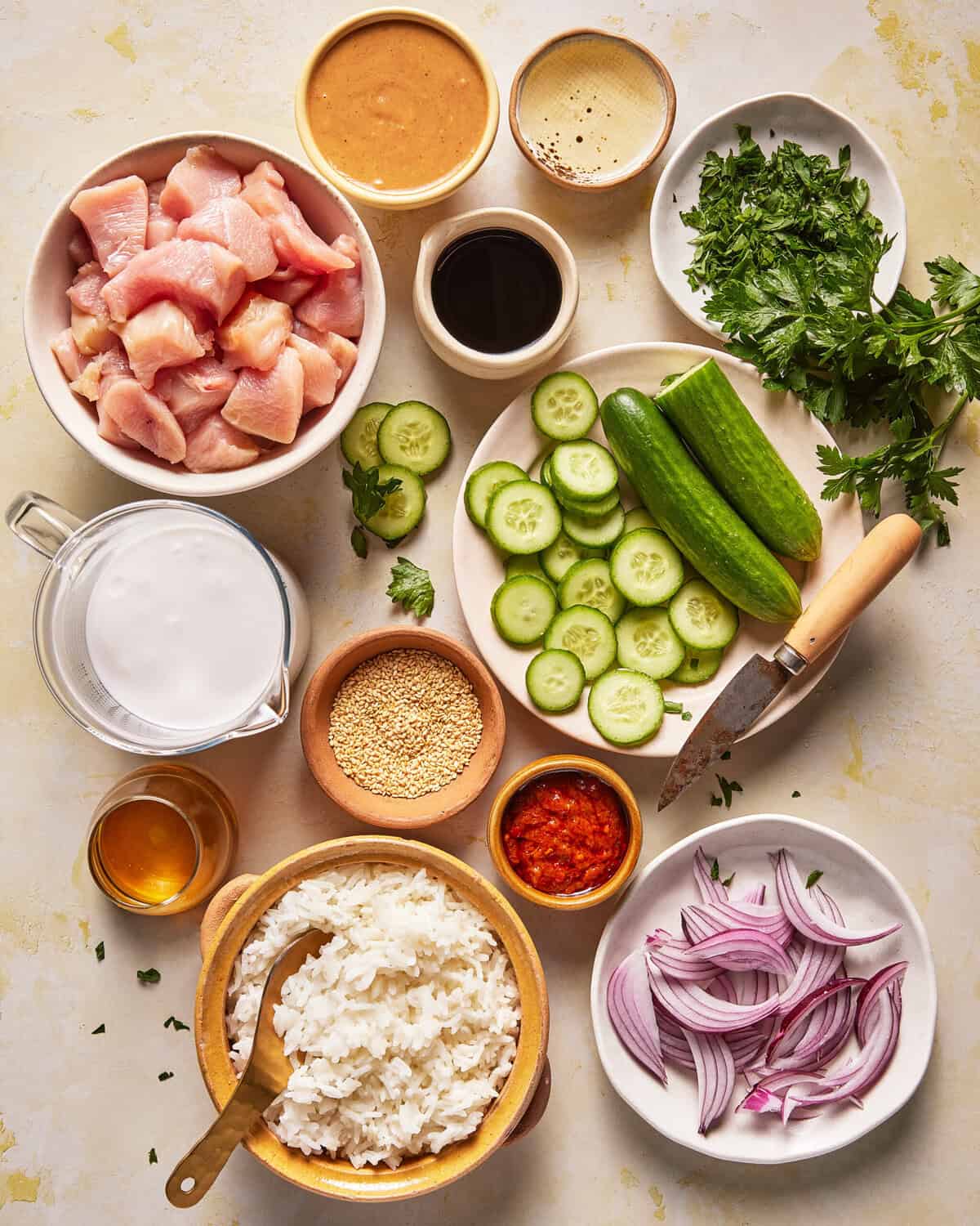 Overhead view of bowls with chopped chicken, white rice, cucumber slices, red onion, parsley, sauces, coconut milk, sliced cucumber, sesame seeds, and a knife on a cutting board on a light surface.