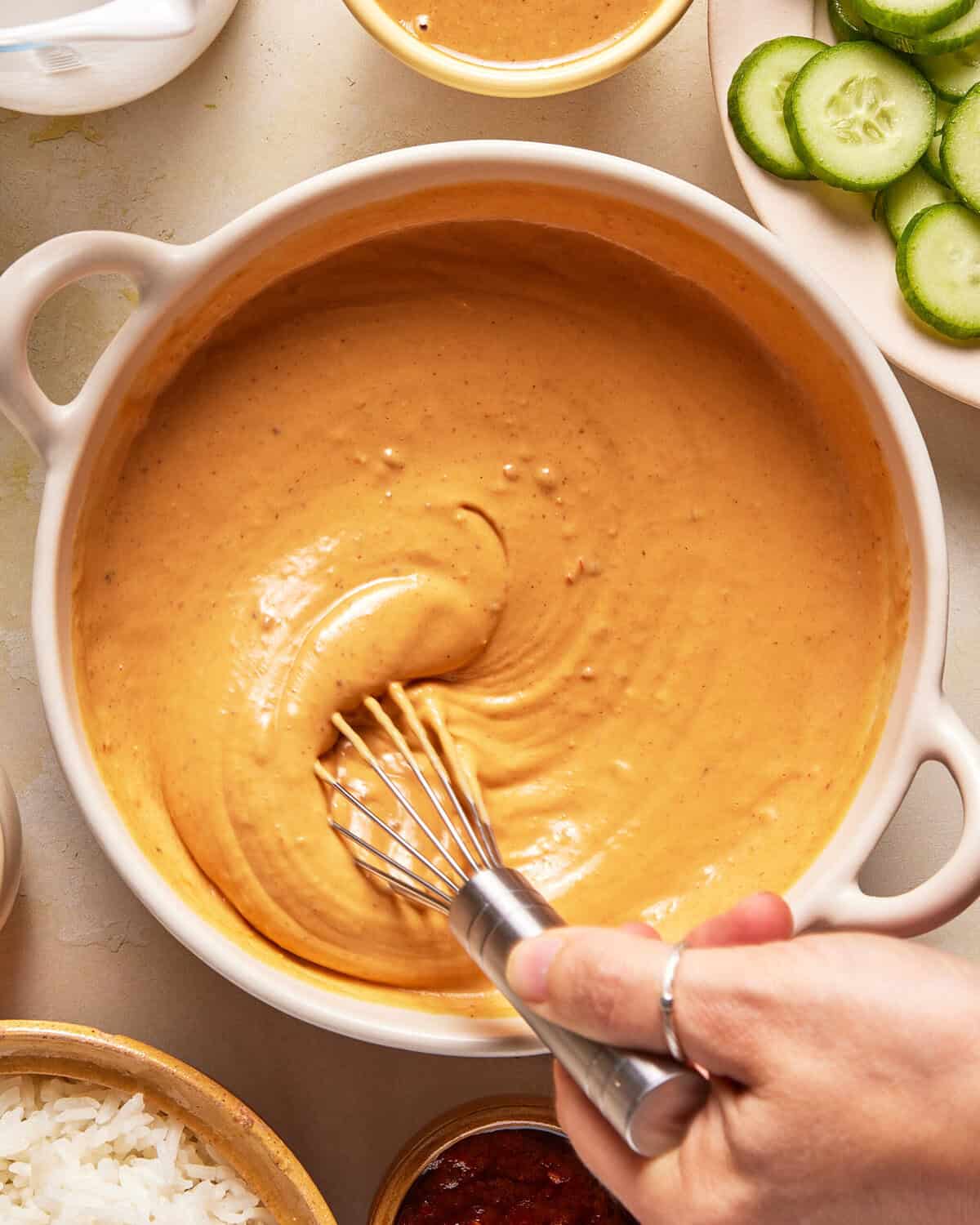 A hand whisks creamy orange sauce in a white bowl, surrounded by sliced cucumbers, cooked rice, and small bowls of condiments.