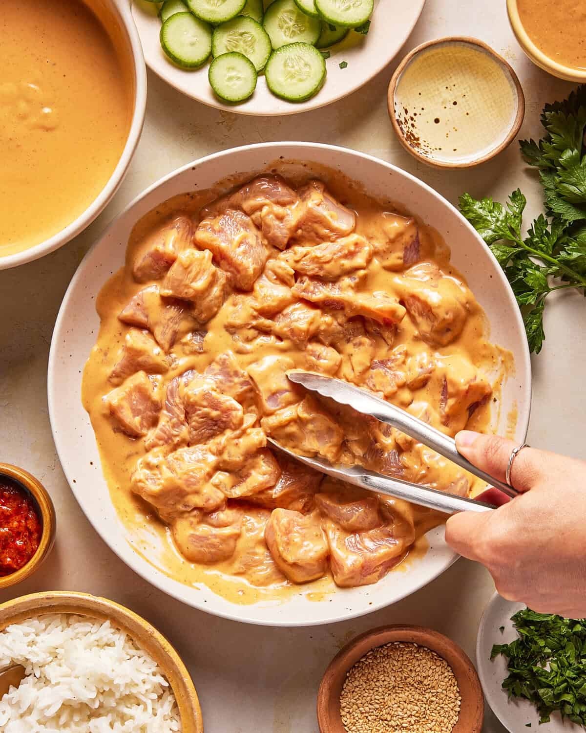 A hand uses tongs to pick up pieces of marinated chicken in a bowl, surrounded by sliced cucumbers, sauce, rice, chili paste, sesame seeds, and fresh herbs on a table.