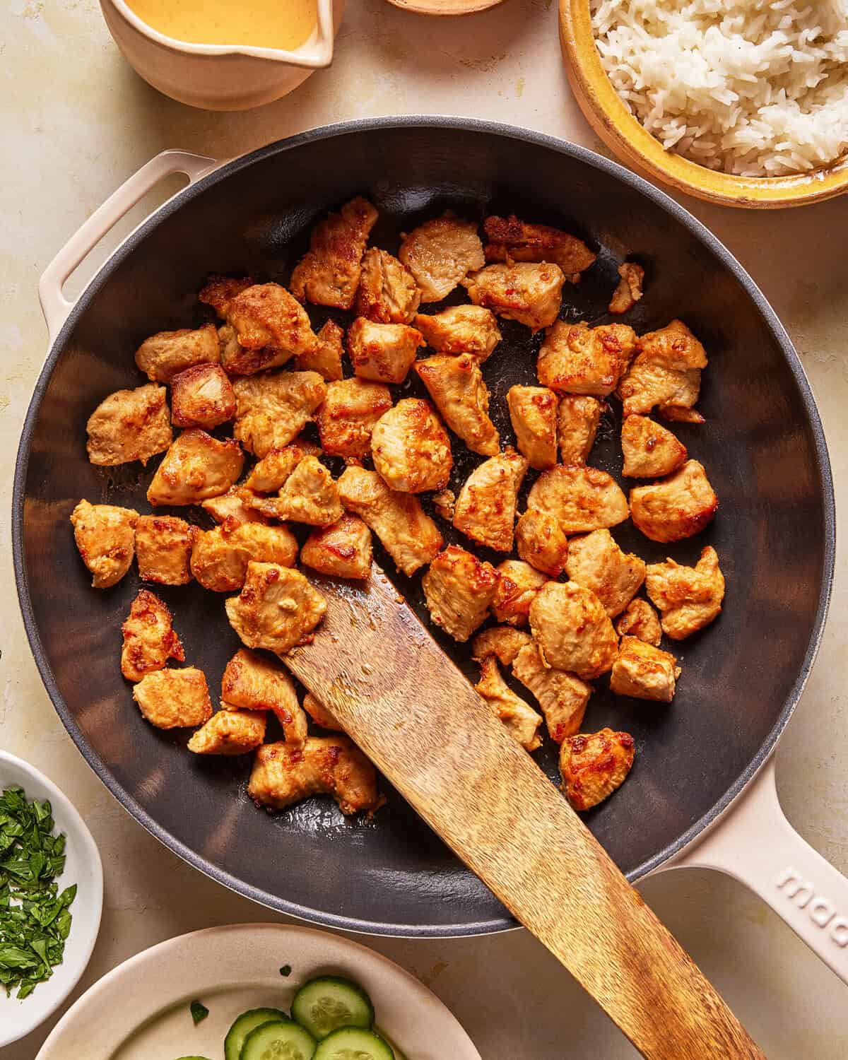 A skillet filled with cooked, seasoned chicken pieces, stirred with a wooden spatula. Surrounding the pan are bowls of white rice, sauce, cilantro, and sliced cucumbers on a light countertop.