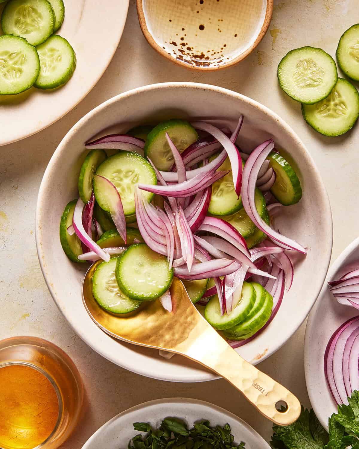 A bowl filled with sliced cucumbers and red onions, with a gold spoon. Surrounding the bowl are plates of cucumber slices, herbs, a small bowl of dressing, and a glass of liquid.