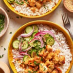 Two yellow bowls of white rice topped with grilled chicken, creamy sauce, sliced cucumbers, red onions, and chopped herbs. Small side bowls with parsley and sesame seeds are nearby, along with a fork.