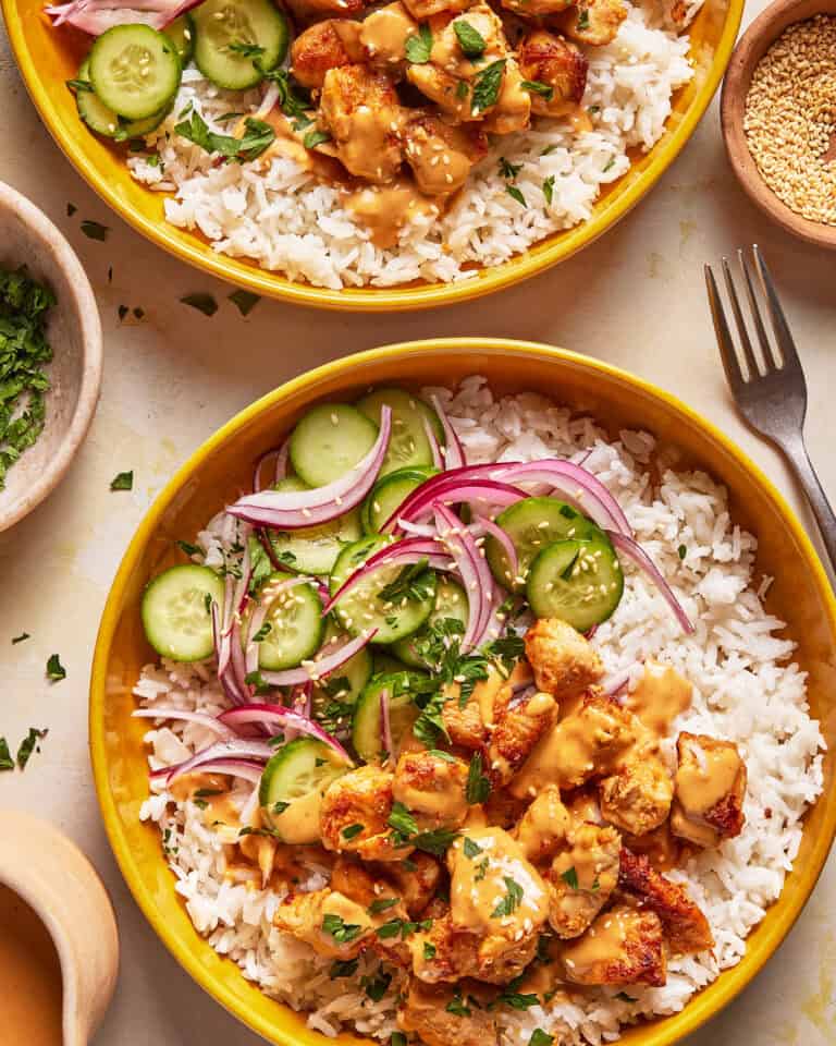 Two yellow bowls of white rice topped with grilled chicken, creamy sauce, sliced cucumbers, red onions, and chopped herbs. Small side bowls with parsley and sesame seeds are nearby, along with a fork.