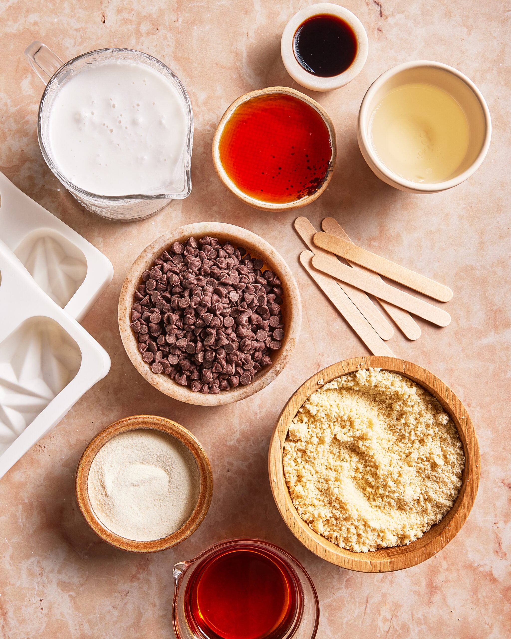 Overhead view of bowls and cups holding chocolate chips, almond flour, sweetener, coconut milk, red liquid, vanilla, and oil, plus wooden sticks and an empty popsicle mold on a beige countertop.