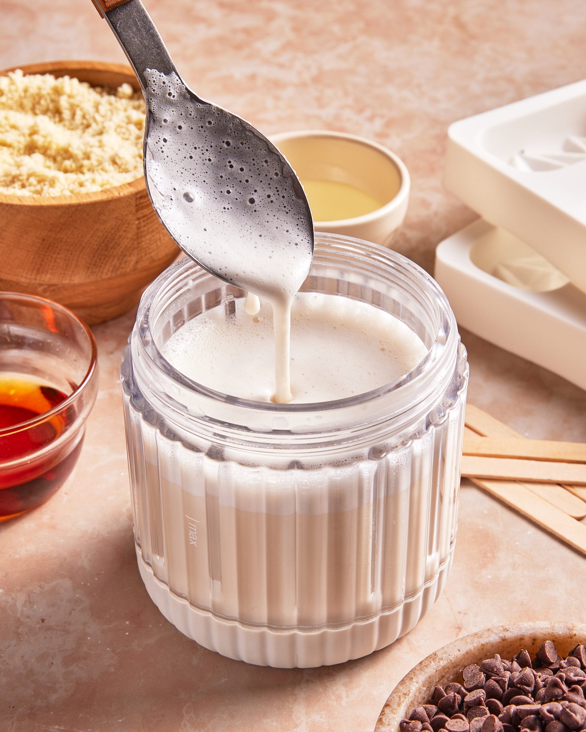 A spoon drips creamy white liquid over a clear container; surrounding are chocolate chips, almond flour, and bowls of oil and syrup, set on a countertop with popsicle molds nearby.